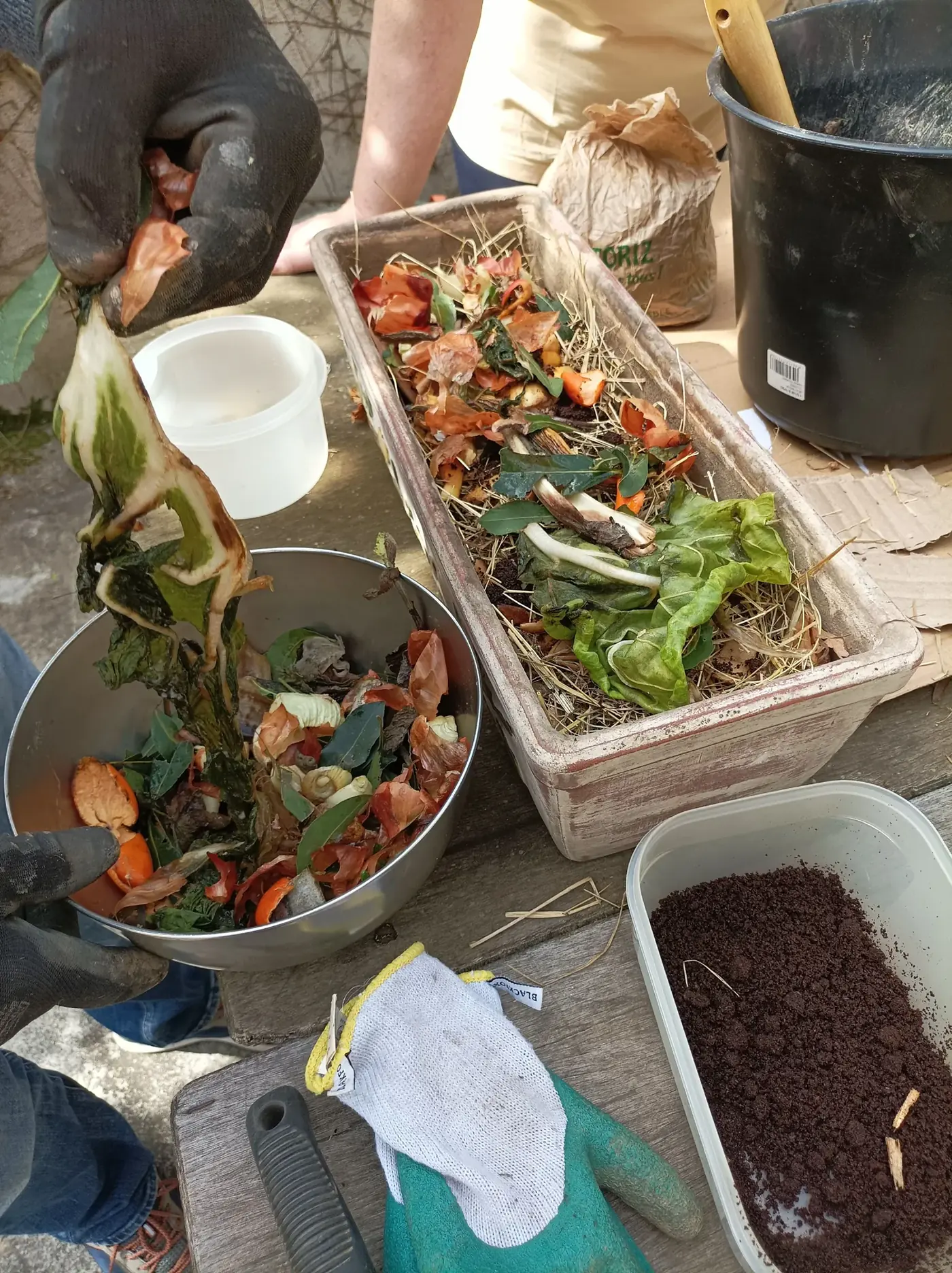 Des mains gantées ajoutent des épluchures à une jardinière pendant l'atelier Tous au compost